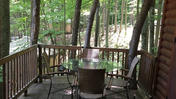 Un espace extérieur abrité, doté d’une table en verre et de chaises en osier, entouré de rampes en bois et d’arbres.