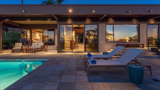 Una casa moderna amb piscina, mobiliari exterior i grans portes de vidre que donen a un interior ben il·luminat.