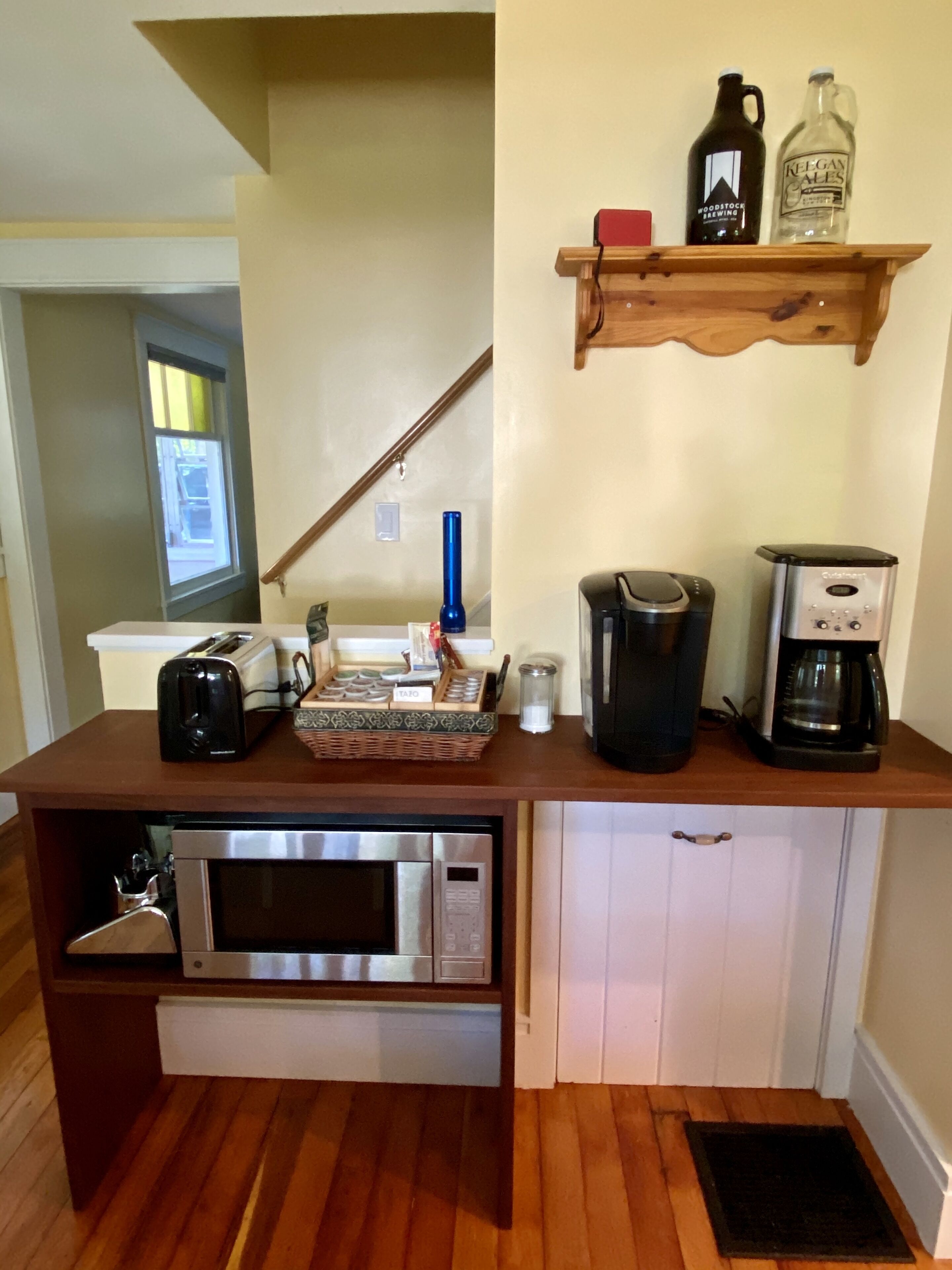 A compact kitchen with a built-in microwave, a coffee maker, and a toaster. There is a wooden shelf above the counter holding a bottle and a jug.