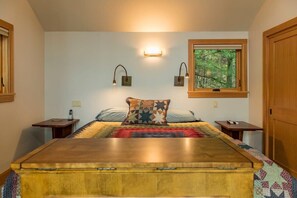 Quarto com uma cama grande, duas mesas de cabeceira, uma janela com vista para árvores e luminárias fixadas na parede.