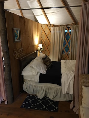 Quarto aconchegante com teto de madeira, cama com roupa de cama branca, mesa de cabeceira com abajur e janela com cortinas.