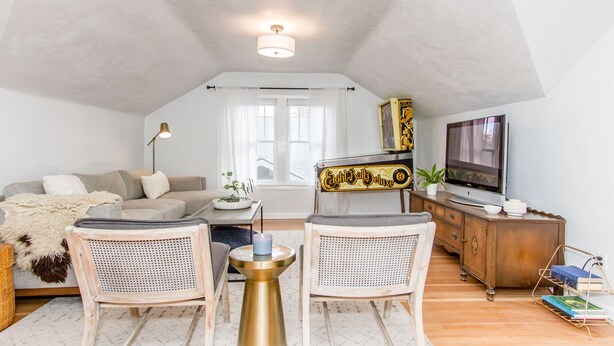 Um apartamento loft aconchegante com teto alto, sofá, duas cadeiras, mesa de apoio, armário, televisão e janela com cortinas.