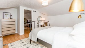 Quarto com cama, cômoda, escrivaninha e cadeira. Há uma luminária no teto e um abajur na parede.