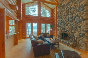 A log cabin interior with a stone fireplace, wooden walls, and a large window overlooking a scenic view. - Spacious Lakefront Log Cabin w/ boat slip (Mccall)