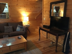 Una acogedora habitación de cabaña de madera con sofá, televisión sobre un mueble bajo, lámpara y ventana con cortinas.