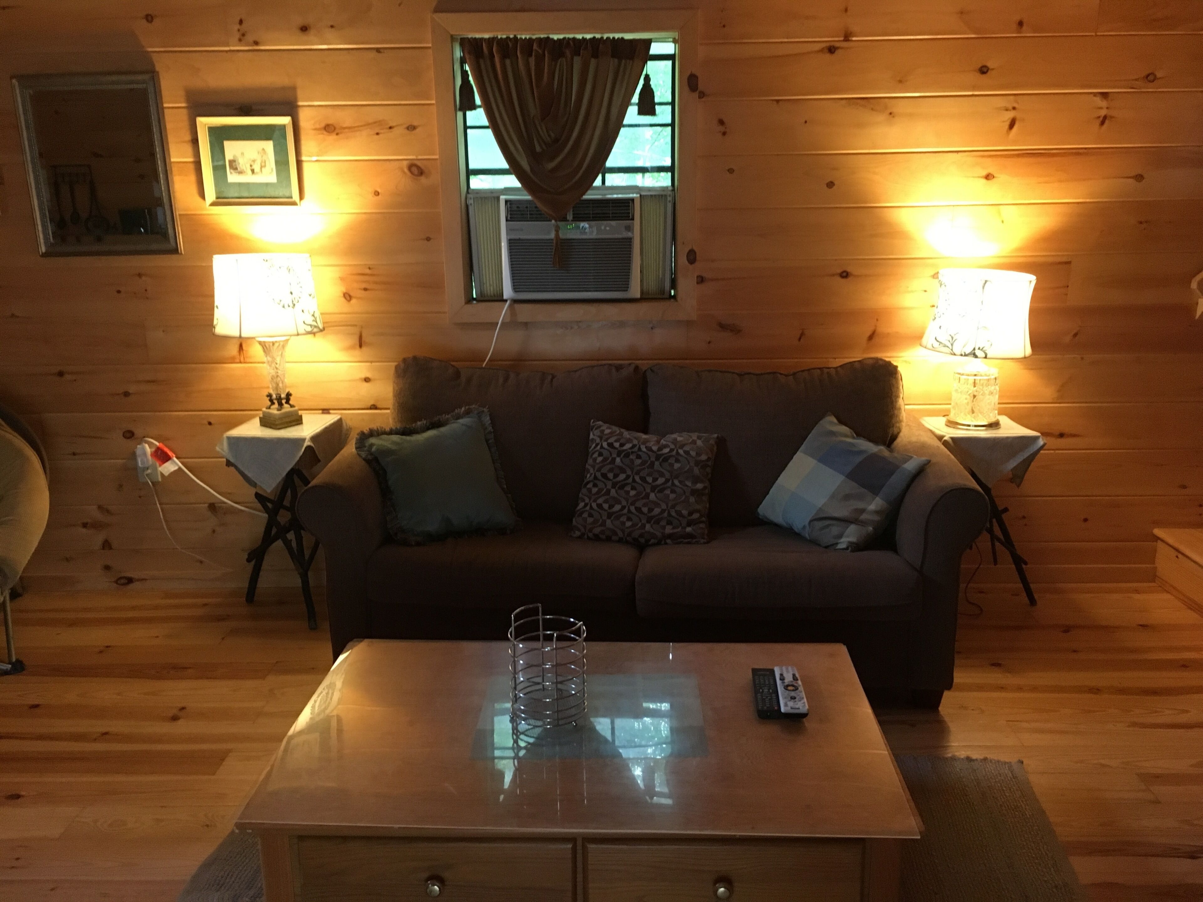 Un salon chaleureux avec un plancher en bois, un canapé marron agrémenté de coussins à motifs, deux petites tables d’appoint avec des lampes et une table basse au centre.