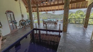 A covered pool area with a wooden ceiling, a dining table with chairs, and a view of a body of water and hills in the background. - Updated Caribbean retreat, with solar and generator power backup (Cruz Bay)