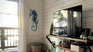 A living room with a TV mounted on a wooden wall, a seahorse painting, a window with curtains, and a wooden floor. - Eternal Summer 2-bedroom villa steps to Coligny Beach and Plaza (Hilton Head Island)