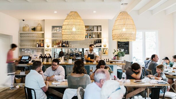 En restaurang med gäster som sitter vid borden, en bardisk med hyllor fulla av flaskor och hängande lampor av rotting.