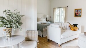 Ett mysigt rum med en säng, en vit soffa, en flätad fåtölj, ett litet bord med glas och en flaska samt en gitarr.