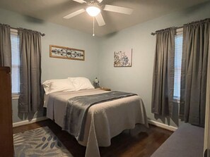 A bedroom with a bed, curtains, a ceiling fan, and artwork on the walls.