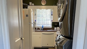 Una cucina compatta con lavastoviglie, frigorifero, fornello e microonde. Si vede una finestra con persiane, un lavello e un piano di lavoro con diversi oggetti.