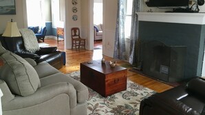 Ein Wohnzimmer mit Kamin, einem darüber montierten Fernseher und einem Sofa. Ein hölzerner Wickelkommode und eine Lampe auf einem Tisch sind ebenfalls vorhanden.