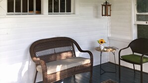 Eine überdachte Veranda mit Deckenventilator, Korbstühle, einem kleinen Tisch und einer Vase mit Sonnenblumen sowie einem Teppich.