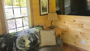 Une chambre chaleureuse dans un chalet, avec un grand couvre-lit orné de motifs d’ours, une petite table avec une lampe et une fenêtre donnant sur la forêt.