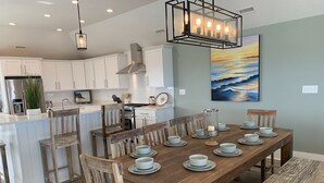 A modern kitchen with white cabinets, a wooden dining table set with plates, and a painting of a sunset on the wall. - Steps to Beach*Private Heated Pool*Elevator* Jennette's Pier (Nags Head)
