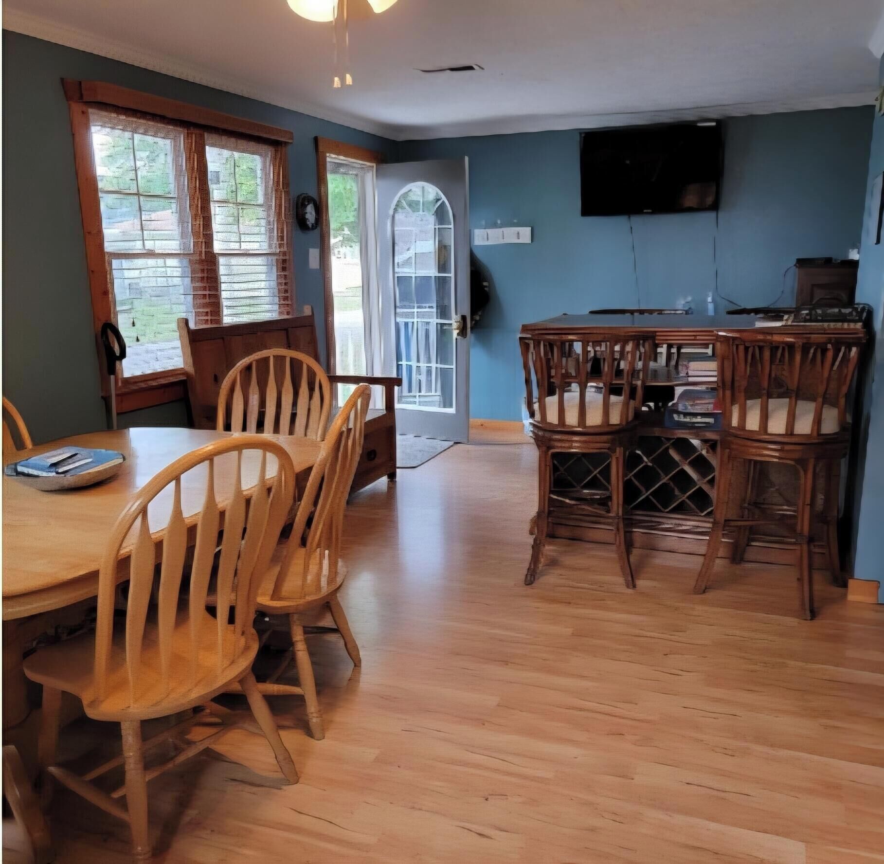 A dining area with wooden chairs and a table, a television mounted on the wall, and a door leading outside.