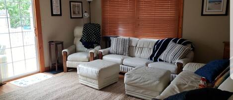 A living room with a beige sofa, a white armchair, and a coffee table. There is a window with blinds, a lamp, and framed pictures on the wall.