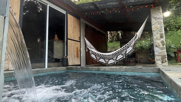 A hot tub with a waterfall feature, a hammock, and a stone structure. - CHARMING  FOREST LOG CABIN (utuado)