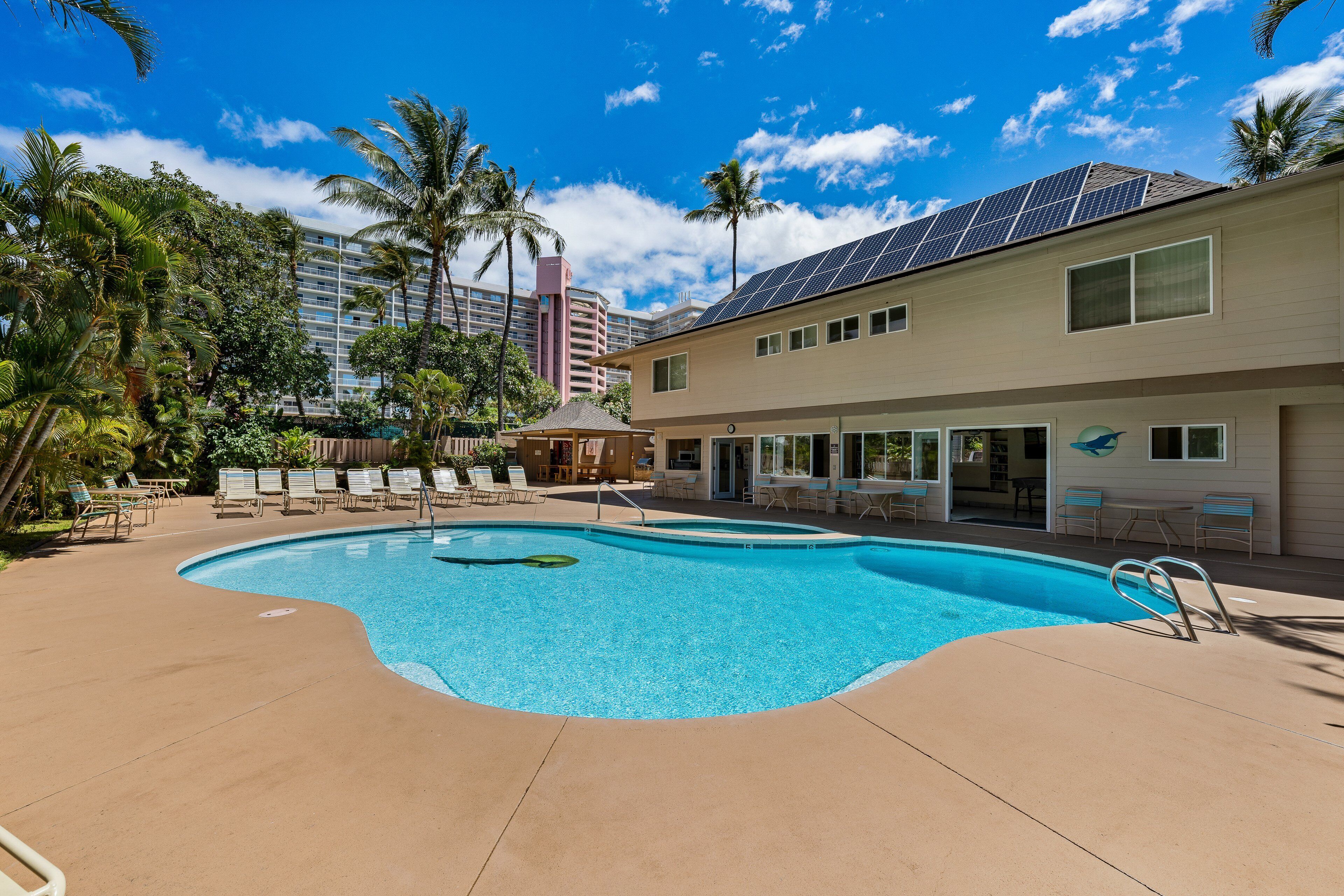 A swimming pool with lounge chairs, a covered seating area, and a building with solar panels.