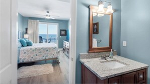 A bedroom with a bed, a ceiling fan, a window with curtains, a dresser, and a bathroom with a sink, mirror, and towel rack.