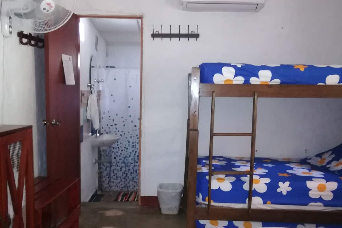 A small, compact room with a bunk bed, a single bed, a wooden wardrobe, a fan, a sink, and a tiled floor.