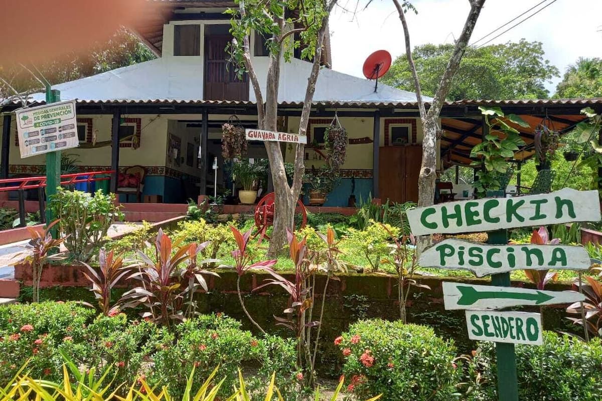 A signpost with directions to 'CHECKIN PISCINA' and 'SENDERO' in a garden area with a building in the background.