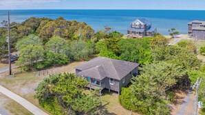 A coastal house with a large deck, surrounded by dense trees and a view of the ocean.