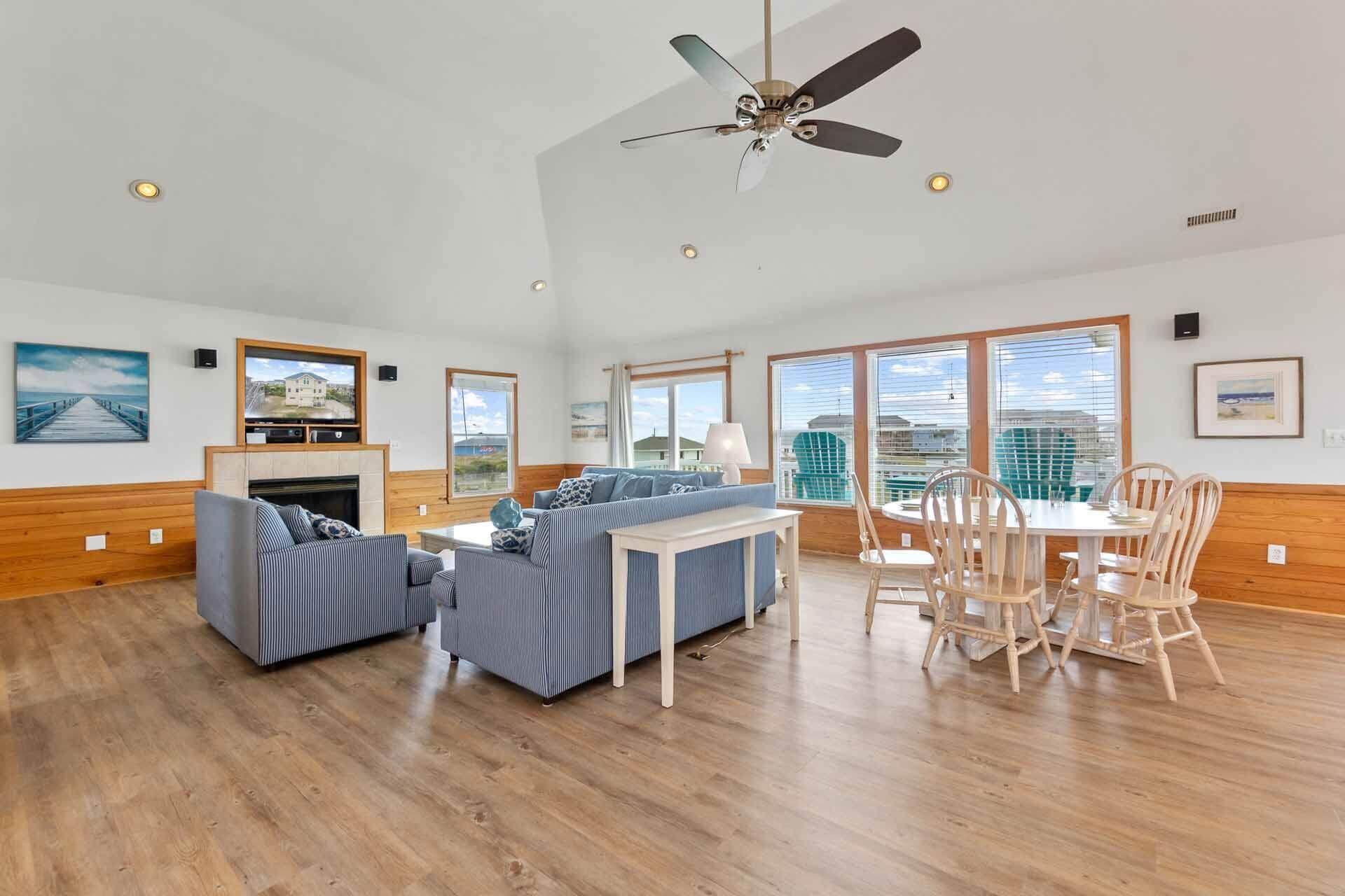 A spacious living room with a fireplace, a ceiling fan, and large windows offering a view of the sea.