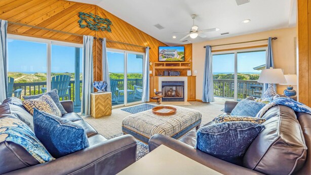 A spacious living room with a fireplace, large windows, and a view of the landscape. - Oceanfront Avon Home with Private Pool, Hot Tub, Deluxe Game Room, Tiki Bar (Avon)