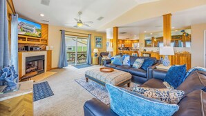 A spacious living room with a fireplace, a large sofa set, a dining area, and a kitchen visible through an open sliding door. - Oceanfront Avon Home with Private Pool, Hot Tub, Deluxe Game Room, Tiki Bar (Avon)
