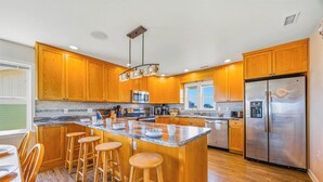 A modern kitchen with wooden cabinets, a central island with stools, stainless steel appliances, and a window providing natural light. - Oceanfront Avon Home with Private Pool, Hot Tub, Deluxe Game Room, Tiki Bar (Avon)
