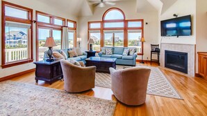 A spacious living room with a fireplace, large windows, and a TV mounted on the wall. - Semi-Oceanfront Home w/ Private Pool, Hot Tub, Elevator, Game Room, and Wet Bar (Salvo)