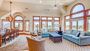 A spacious living room with a sectional sofa, a dining area with a table and chairs, and large windows offering views of the surrounding area. - Semi-Oceanfront Home w/ Private Pool, Hot Tub, Elevator, Game Room, and Wet Bar (Salvo)