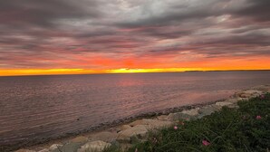 A coastal landscape with a sunset over the water, rocky shoreline, and vegetation.