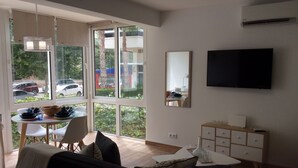 Una sala de estar con un sofá, una mesa de comedor con sillas, un televisor montado en la pared y ventanas amplias con vista a la calle.