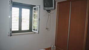 Un dormitorio con ventana, televisor montado en la pared, armario y mesita de noche.