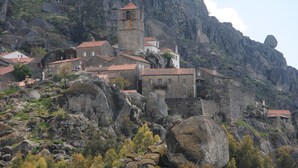 Un edificio de piedra histórico con un campanario, ubicado en una ladera rocosa rodeado de casas.