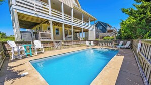A rectangular swimming pool with a wooden deck surrounding it, a covered outdoor seating area, and a view of a residential building in the background. - Oceanview Home with Pool, Hot Tub, Game and Media Rooms. I Love Beach Music! (Salvo)