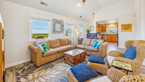 A spacious living room with a large sectional sofa, a coffee table, and wicker chairs. There is a kitchen in the background with wooden cabinets and a window. - Oceanview Home with Pool, Hot Tub, Game and Media Rooms. I Love Beach Music! (Salvo)
