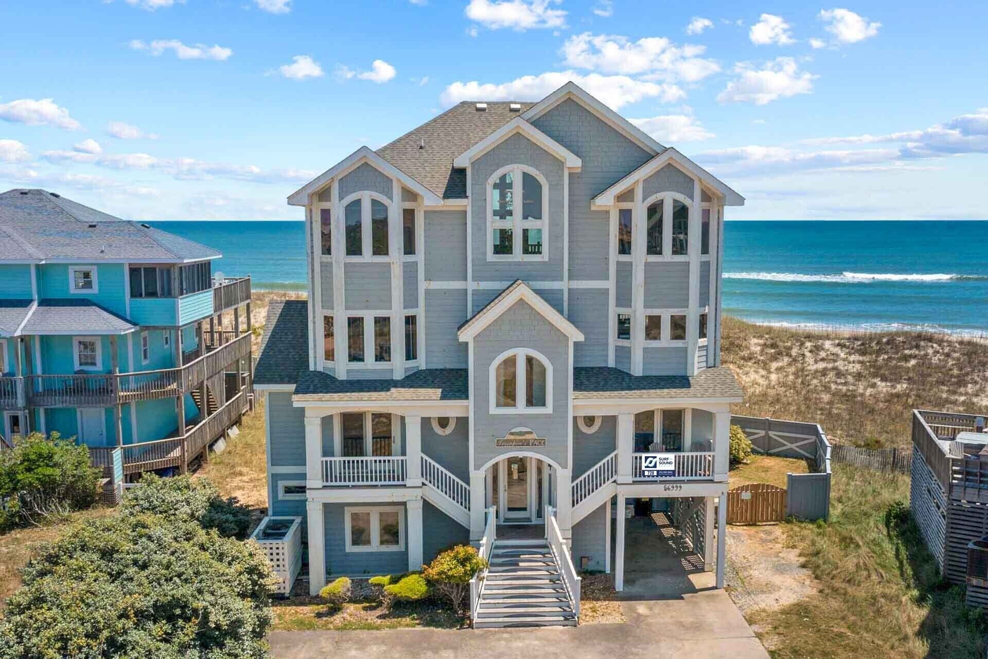 A large, multi-story beachfront house with a prominent front entrance, a covered porch, and a visible beach view.