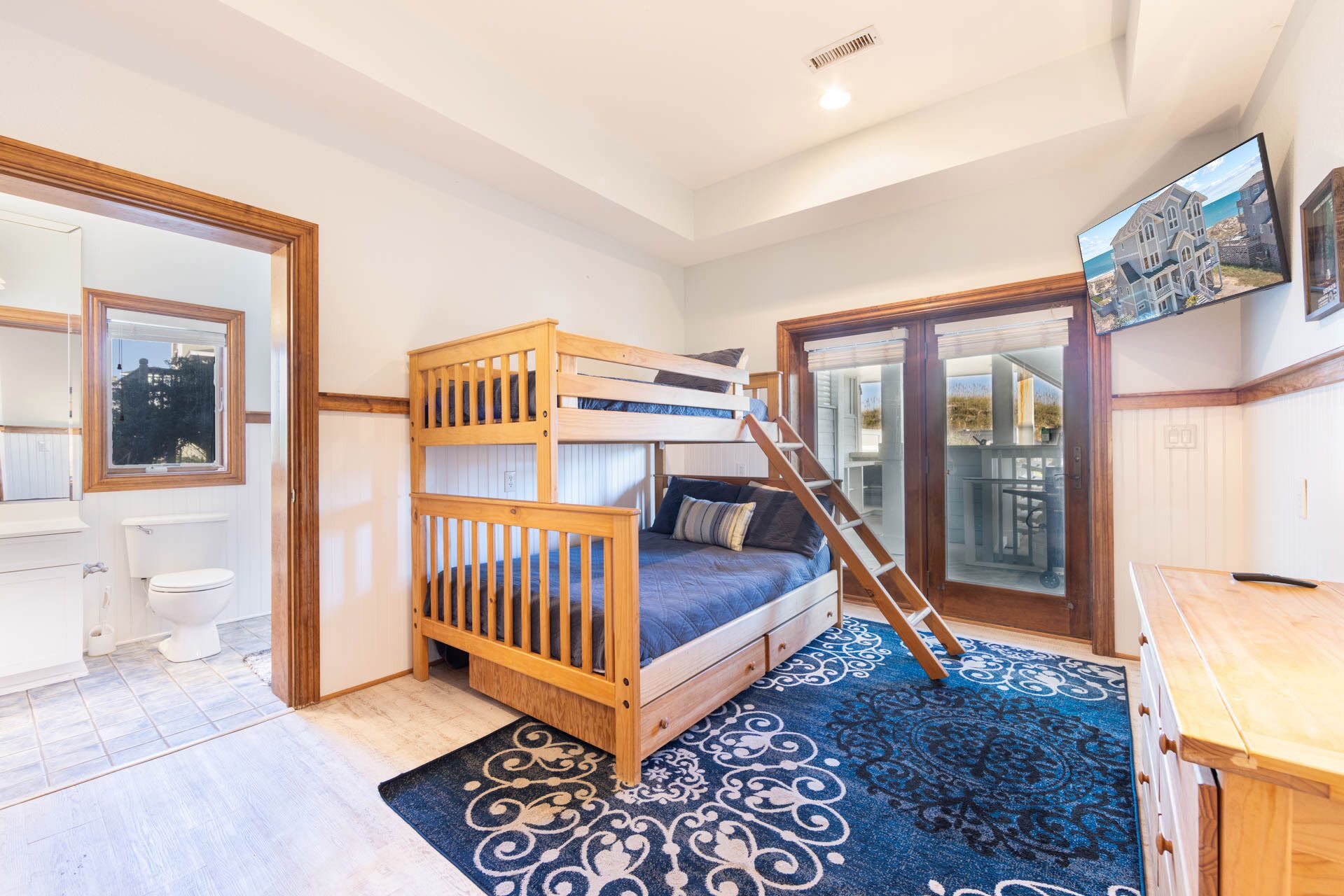 A room with a bunk bed, a wooden desk, a bathroom, and a large window.