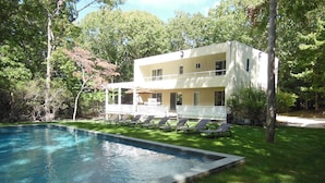 A two-story house with a swimming pool, surrounded by trees and a lawn with sun loungers.