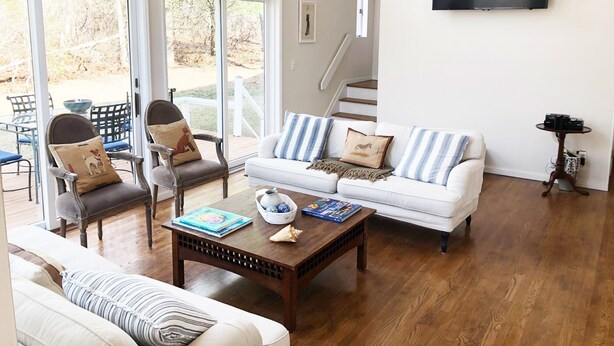 A spacious living room with a white sofa, a wooden coffee table with books, and a TV mounted on the wall.