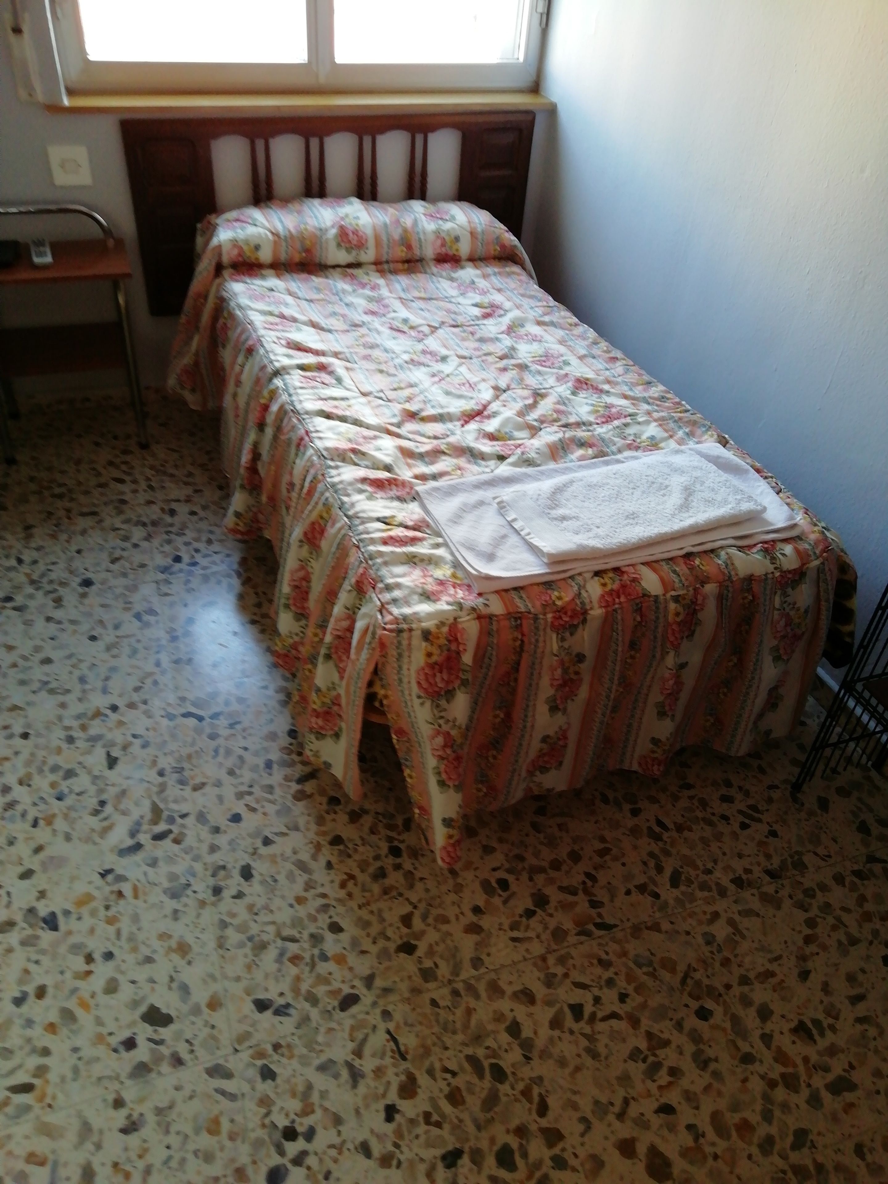 A small, single-bed room with a floral bedspread and a white towel on the bed. The floor is tiled with a speckled pattern.