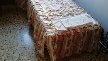 A small, single-bed room with a floral bedspread and a white towel on the bed. The floor is tiled with a speckled pattern.