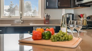 Un piano di lavoro in cucina con un tagliere che contiene peperoni rossi, verdi e gialli, un grappolo d'uva verde, una bottiglia di vino bianco e due bicchieri da vino.