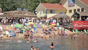 Escena de playa con gente nadando y tomando el sol. Hay sillas y sombrillas de colores, un bar y un edificio con un letrero que dice 'Captain's Bar & Grill'.