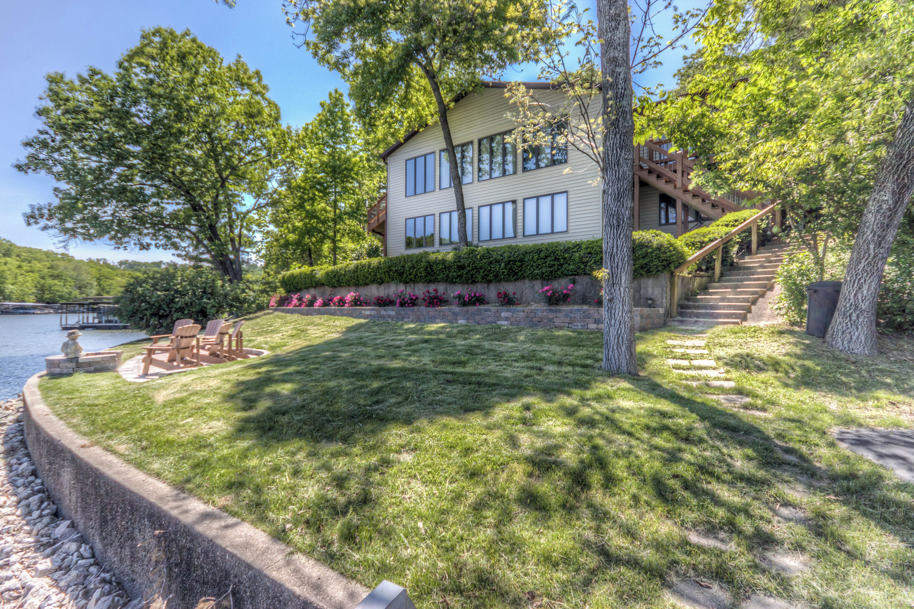 A two-story house with a stone staircase leading to a deck, surrounded by lush greenery and a body of water.