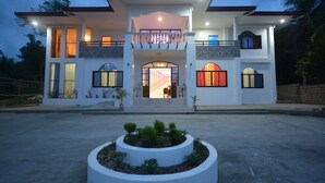 A two-story house with a central entrance, a balcony on the upper floor, and a circular garden in the foreground. - White Summerville Tourist Inn (Busuanga)
